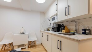 Cocina compacta con microondas, cafetera y pileta. Hay dos sillas blancas y una mesita con lámpara.