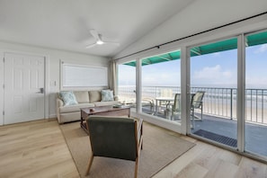 A room with a sofa, a chair, a table, a ceiling fan, and large sliding glass doors leading to a balcony with a view of the ocean.