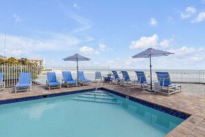 A pool area with blue lounge chairs and striped umbrellas, a clear sky, and a white fence.