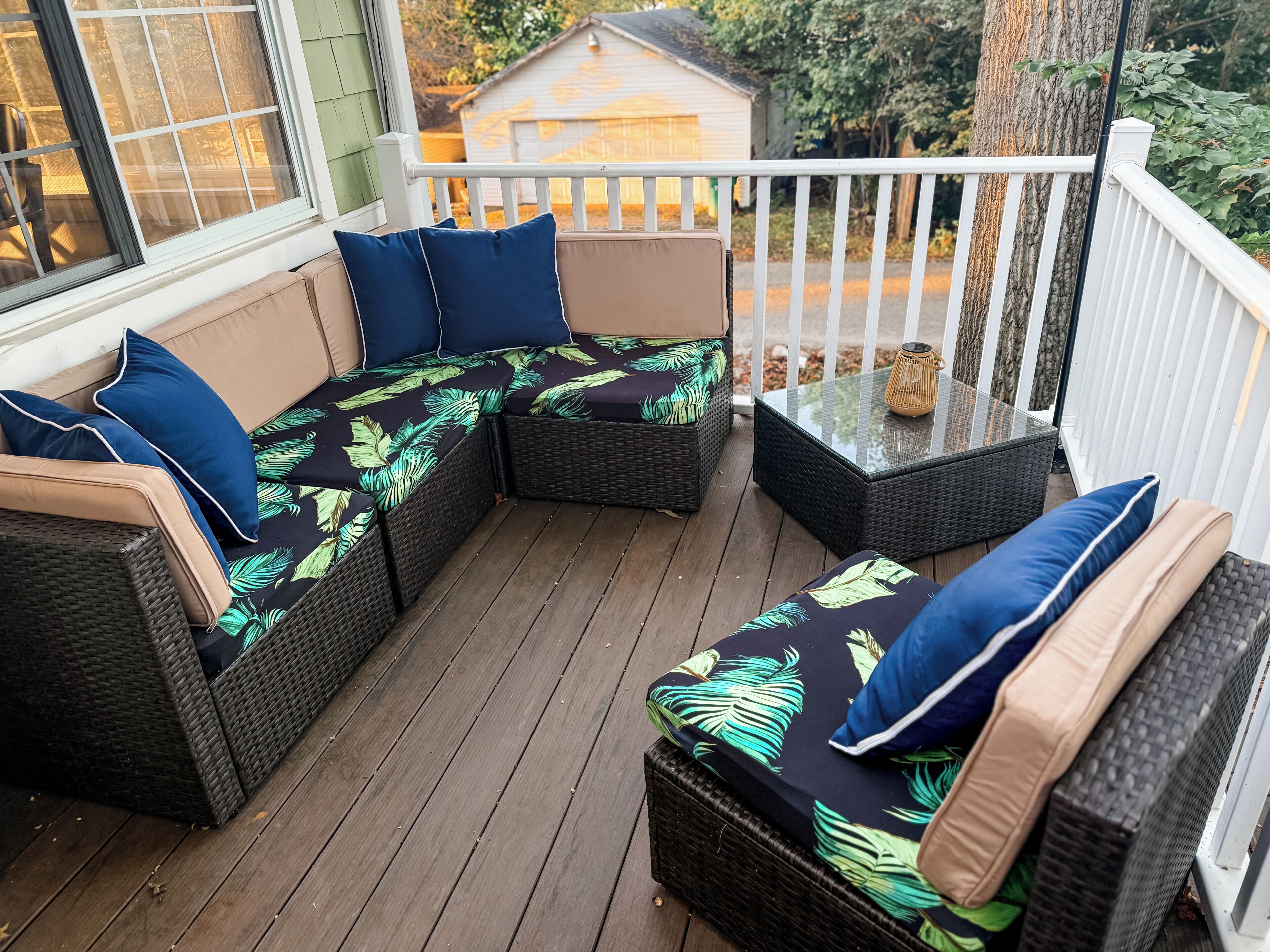 A covered outdoor seating area with wicker furniture and cushions, a glass table, and a view of the surrounding yard.