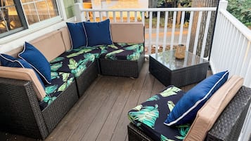 A covered outdoor seating area with wicker furniture and cushions, a glass table, and a view of the surrounding yard.