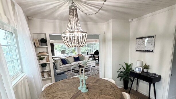 Ein Essbereich mit einem runden Holztisch, weißen Stühlen, einer hängenden Chandelüster und Blick auf ein Wohnzimmer mit einer Couch und einem Esstisch.