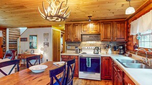 A rustic cabin interior with wooden walls, a dining area with a wooden table and chairs, a kitchen with wooden cabinets and a stove, and a chandelier with antlers.