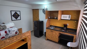 Een compact keukenhoekje met een magnetron, koffiezetapparaat en een kleine koelkast. Er is een houten plank met handdoeken en een lijst met een foto aan de muur.