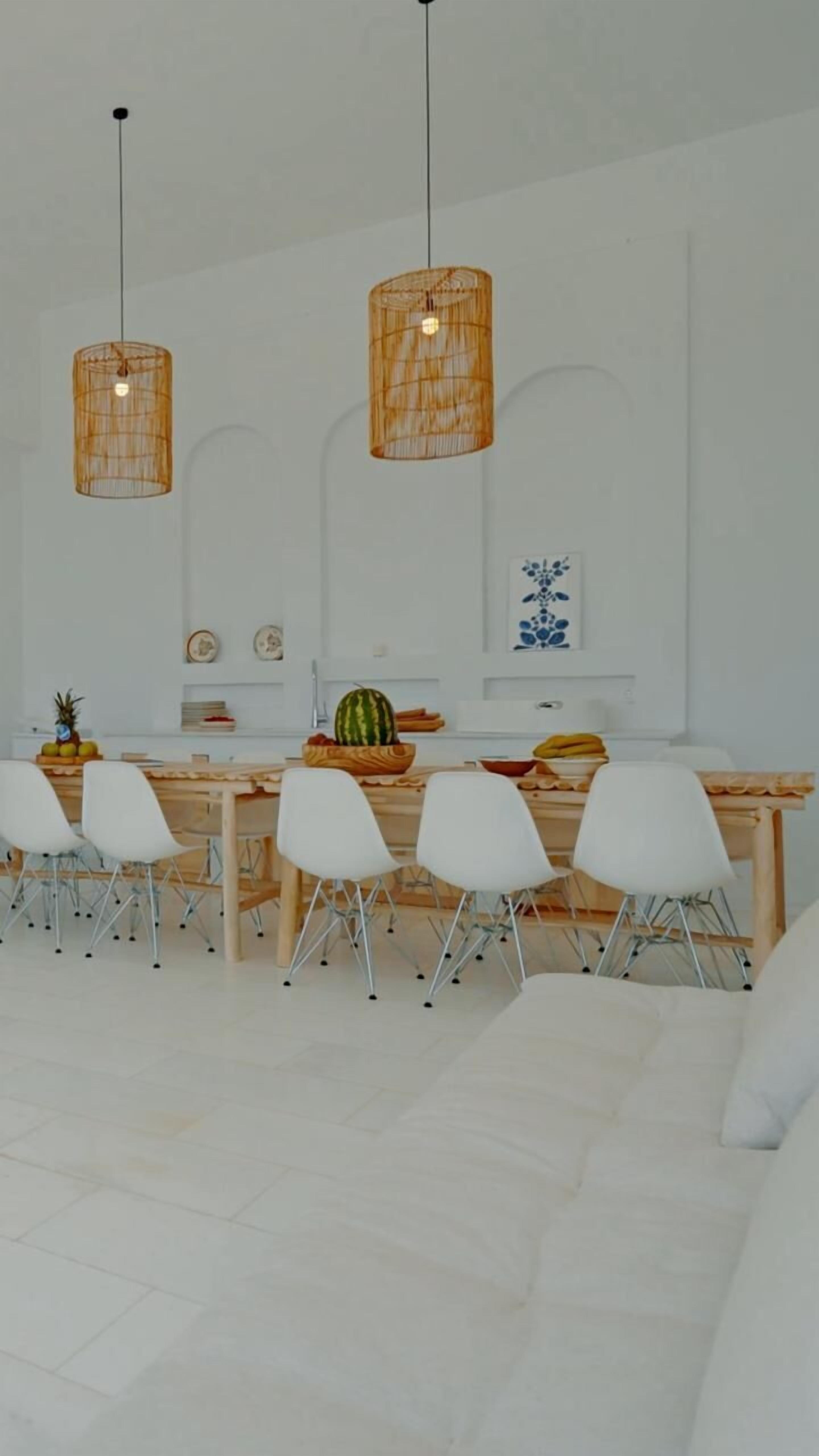 A modern dining area with a long wooden table, white chairs, and two hanging wicker pendant lights.