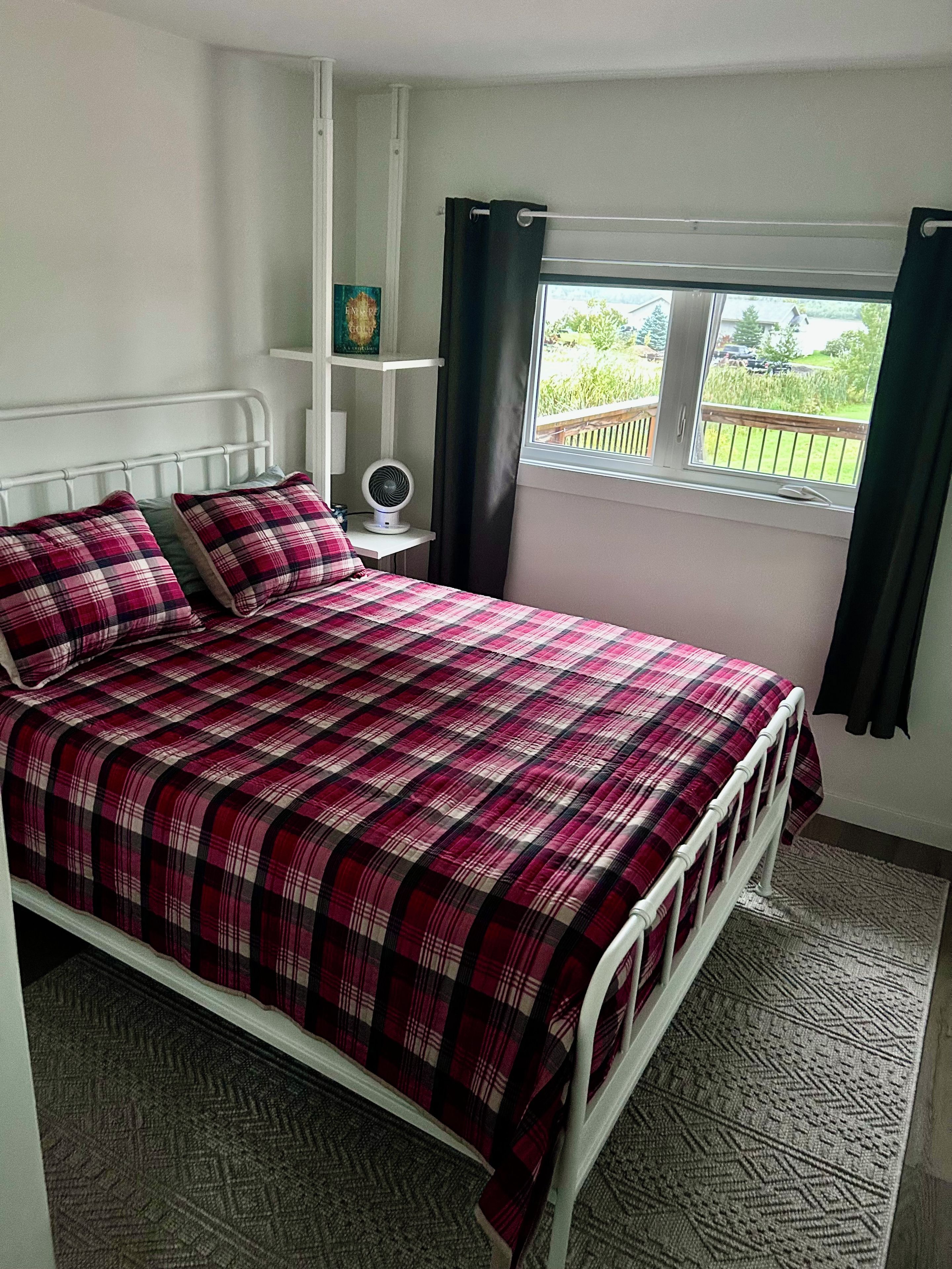 Een slaapkamer met een bed, nachtkastje, ventilator en raam met gordijnen.