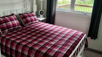 Een slaapkamer met een bed, nachtkastje, ventilator en raam met gordijnen.