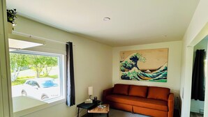 A modern living room with a sofa, a coffee table, and a chair. There is a painting on the wall, a window with a view of a car, and a radiator under the window.
