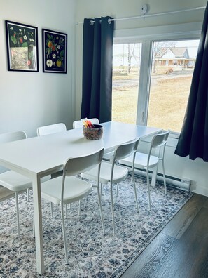 A dining area with a white table and chairs, a patterned rug, and a window with curtains.