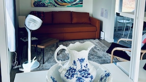 A room with a white table, a white pitcher on a saucer, a red sofa, a standing fan, and a wall-mounted painting.