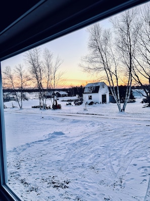 A snowy landscape with a white building and bare trees, viewed through a window. - Coastal Charm Meets Country Calm | Relaxing Cottage with Water Views & Hot Tub (Cocagne)