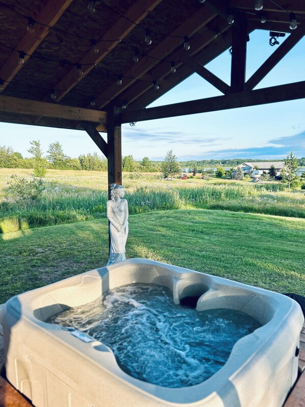 A hot tub under a wooden pergola with a view of a grassy field and trees.