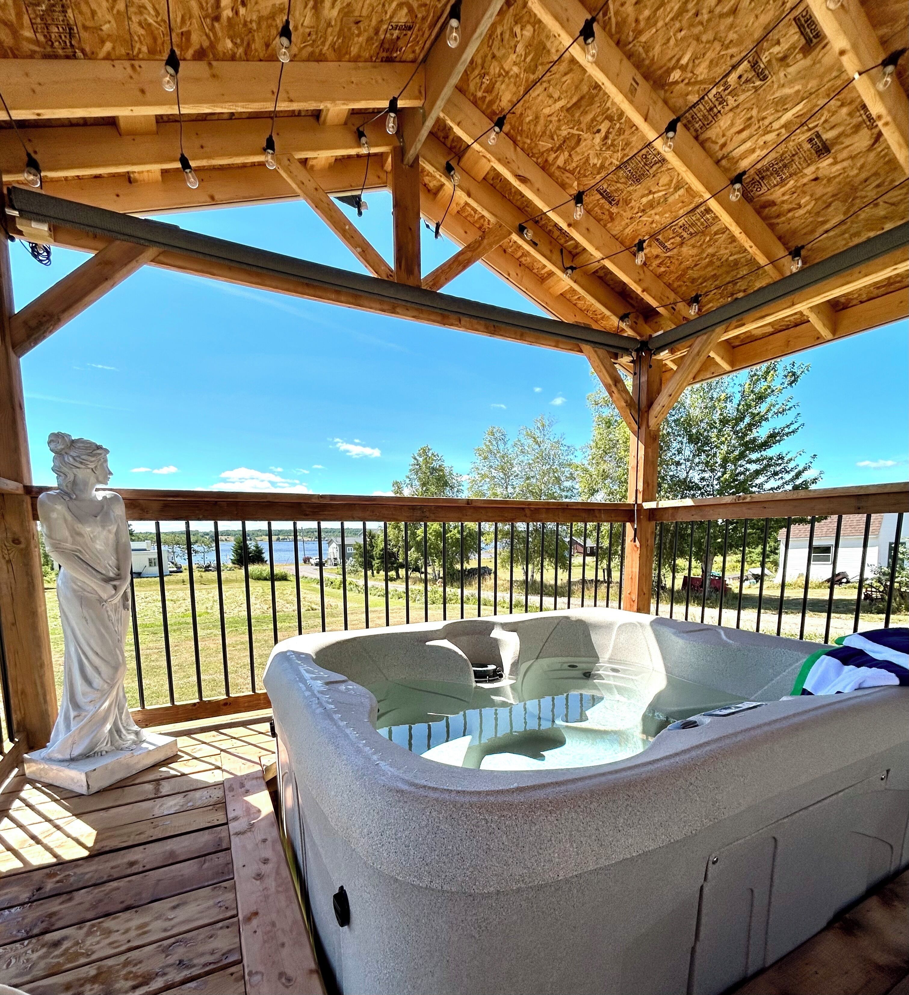 A hot tub under a wooden pergola with a clear view of a lake and a statue.
