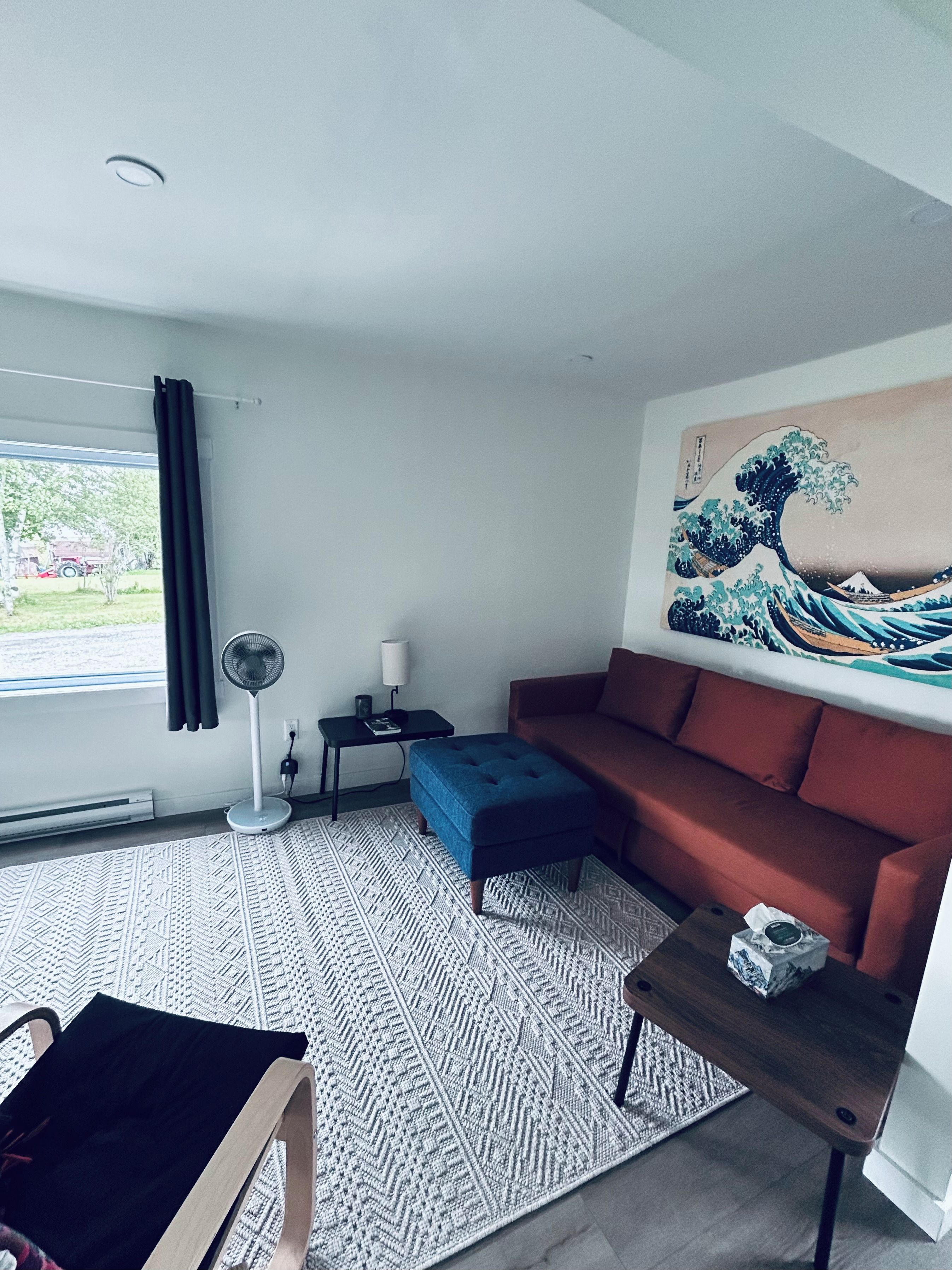 A modern living room with a sofa, ottoman, and a fan. There's a painting on the wall and a window with curtains.