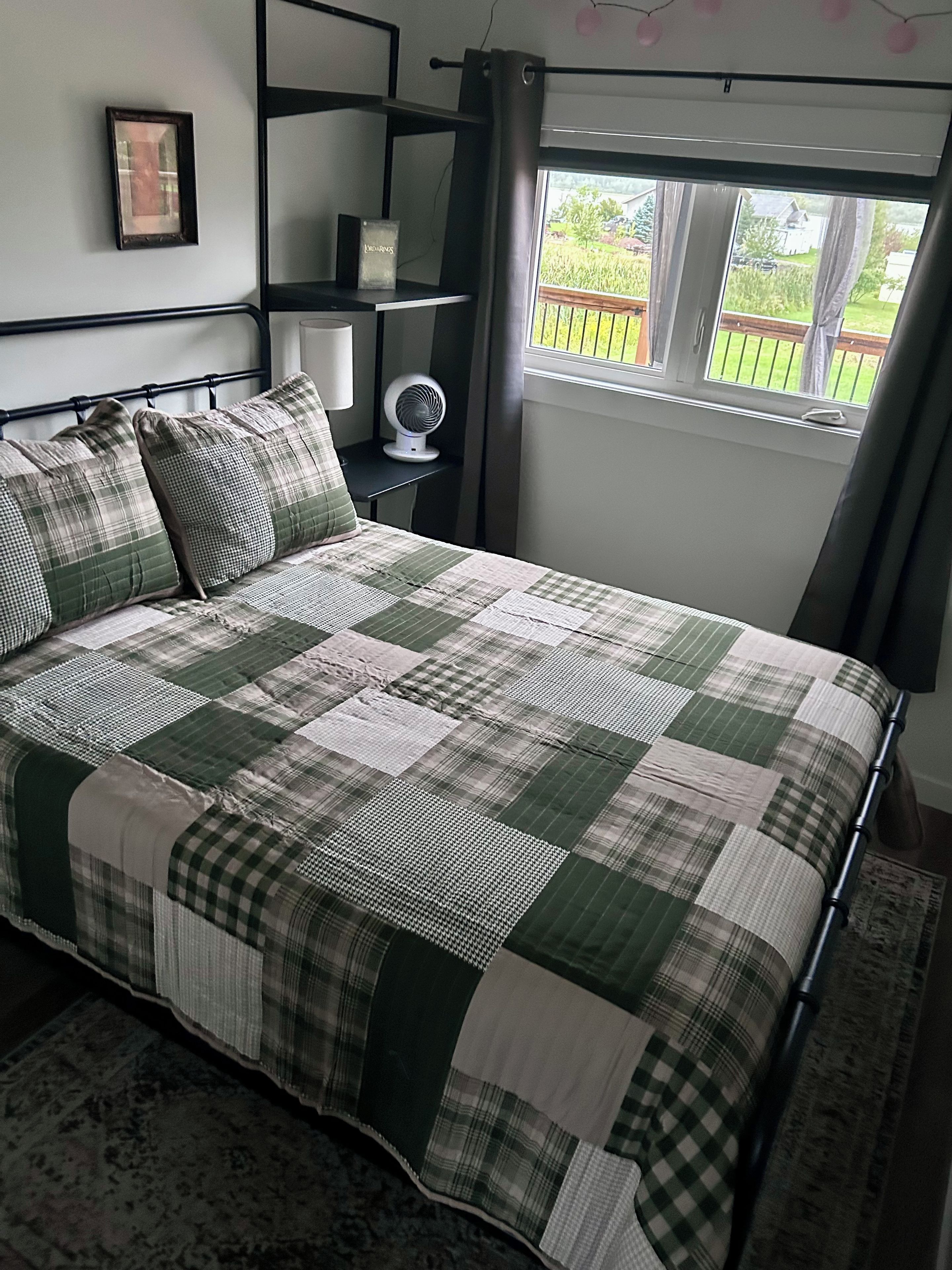A bed with a checkered bedspread, a bedside table with a fan, and a window with a view of a garden.