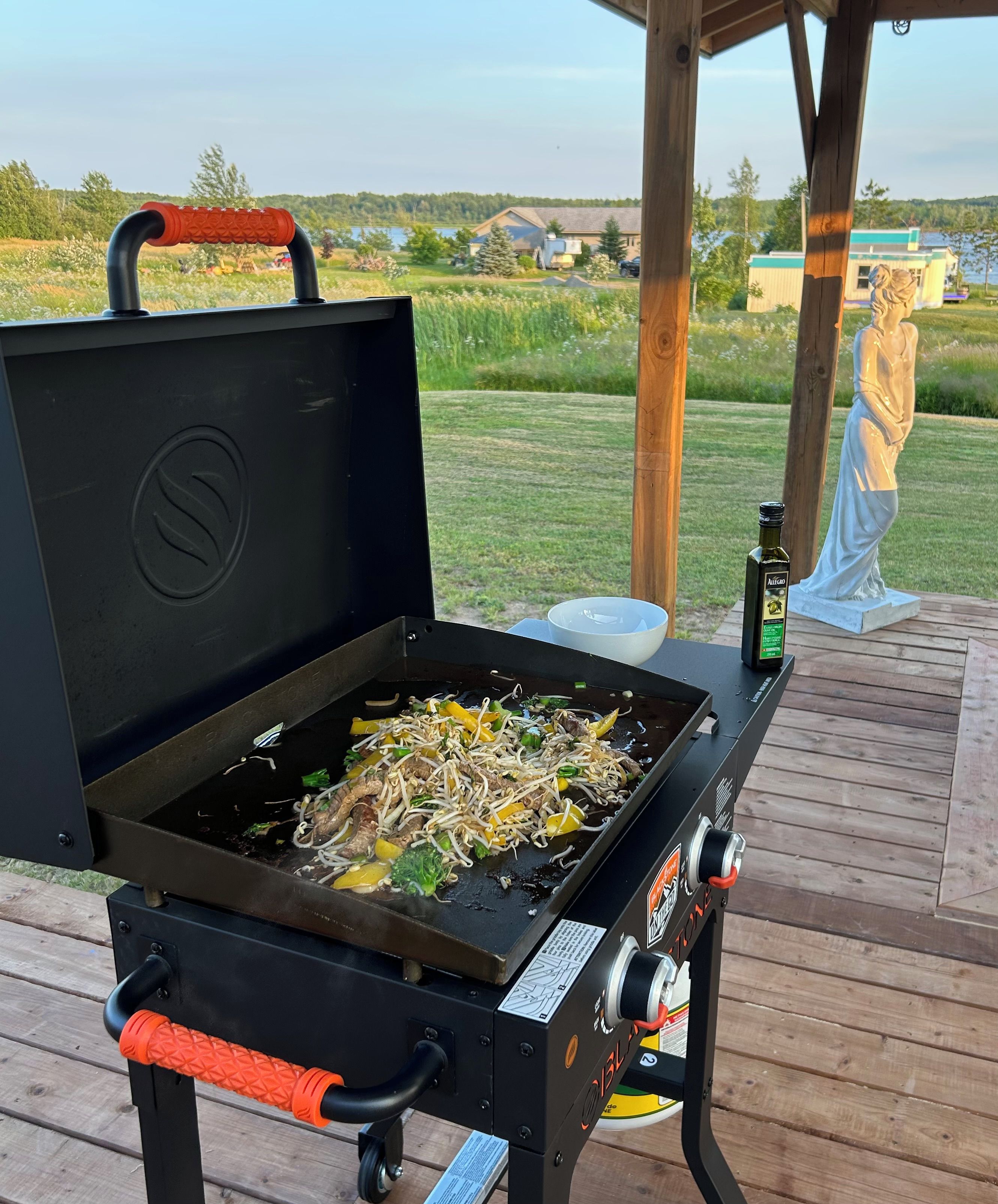 An outdoor grill with food, a bottle, and a statue on a wooden deck.
