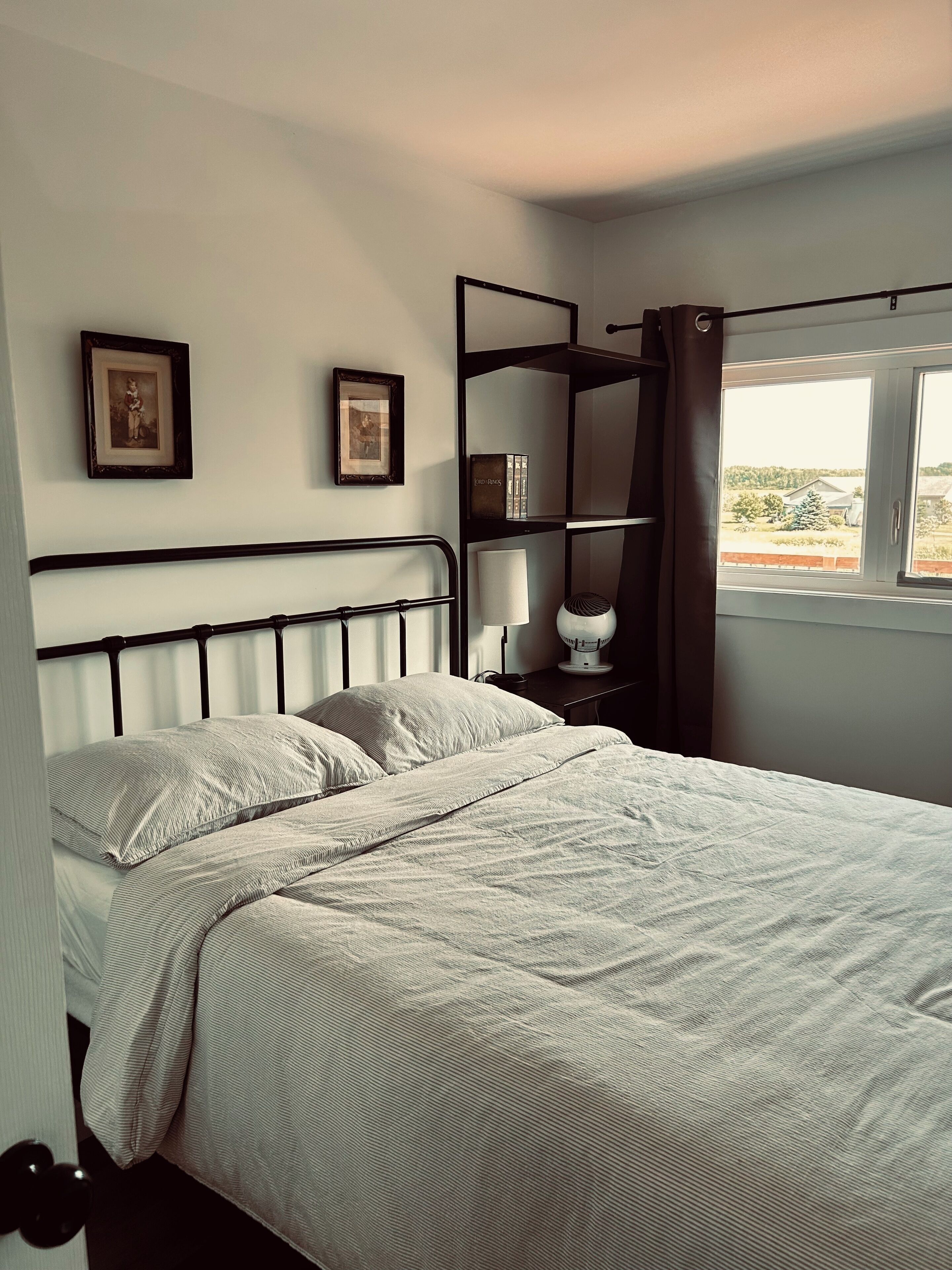 A bedroom with a bed, two framed pictures on the wall, a bedside table with a lamp, and a window with curtains.