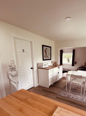 A room with a wooden dining table, white cabinets, a microwave, a framed picture, and a statue. - Coastal Charm Meets Country Calm | Relaxing Cottage with Water Views & Hot Tub (Cocagne)