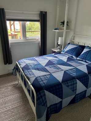 A bedroom with a metal bed frame, a blue and white checkered bedspread, a bedside table with a lamp, and a window with curtains. - Coastal Charm Meets Country Calm | Relaxing Cottage with Water Views & Hot Tub (Cocagne)