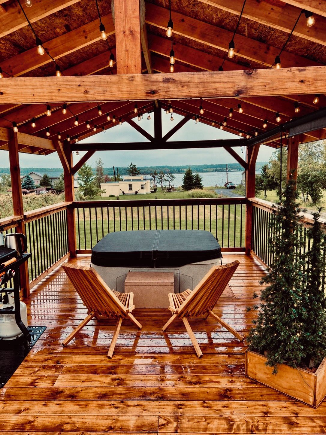 A covered outdoor seating area with a wooden deck, a table with chairs, and a view of a lake and buildings in the distance.