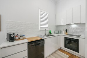 A modern kitchen with white cabinetry, a dishwasher, an oven, and a window with blinds.