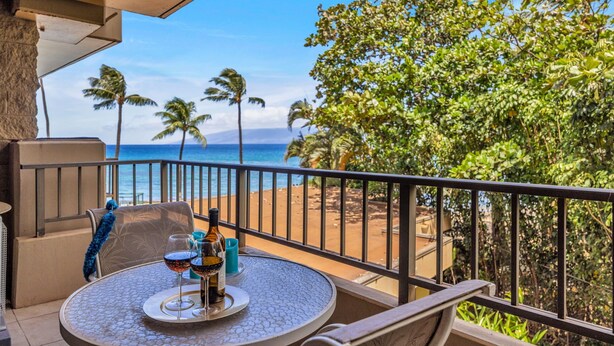 Un balcón con una mesa dispuesta para dos personas, con vista a una playa con palmeras y el océano.