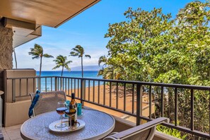 A balcony with a table set for two, overlooking a beach with palm trees and the ocean. - Fully Renovated Condo right on the beach with A/C, Wi-Fi and Everything you need (Lahaina)