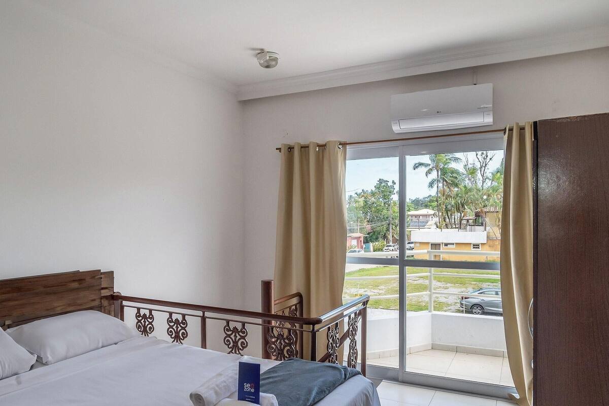 Ein Hotelzimmer mit Bett, Fenster mit Vorhängen und Blick nach draußen, sowie einer Klimaanlage an der Wand.