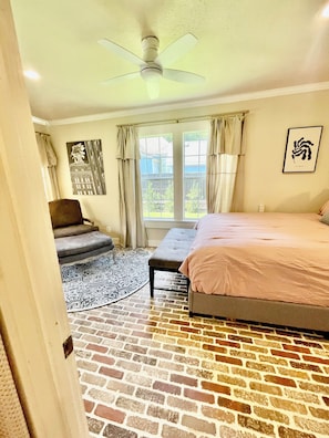 A bedroom with a brick-patterned floor, a bed, a bench, a chair, and a ceiling fan. - Charming 3-bedroom cottage in Historic McKinney, three blocks from the square. (McKinney)