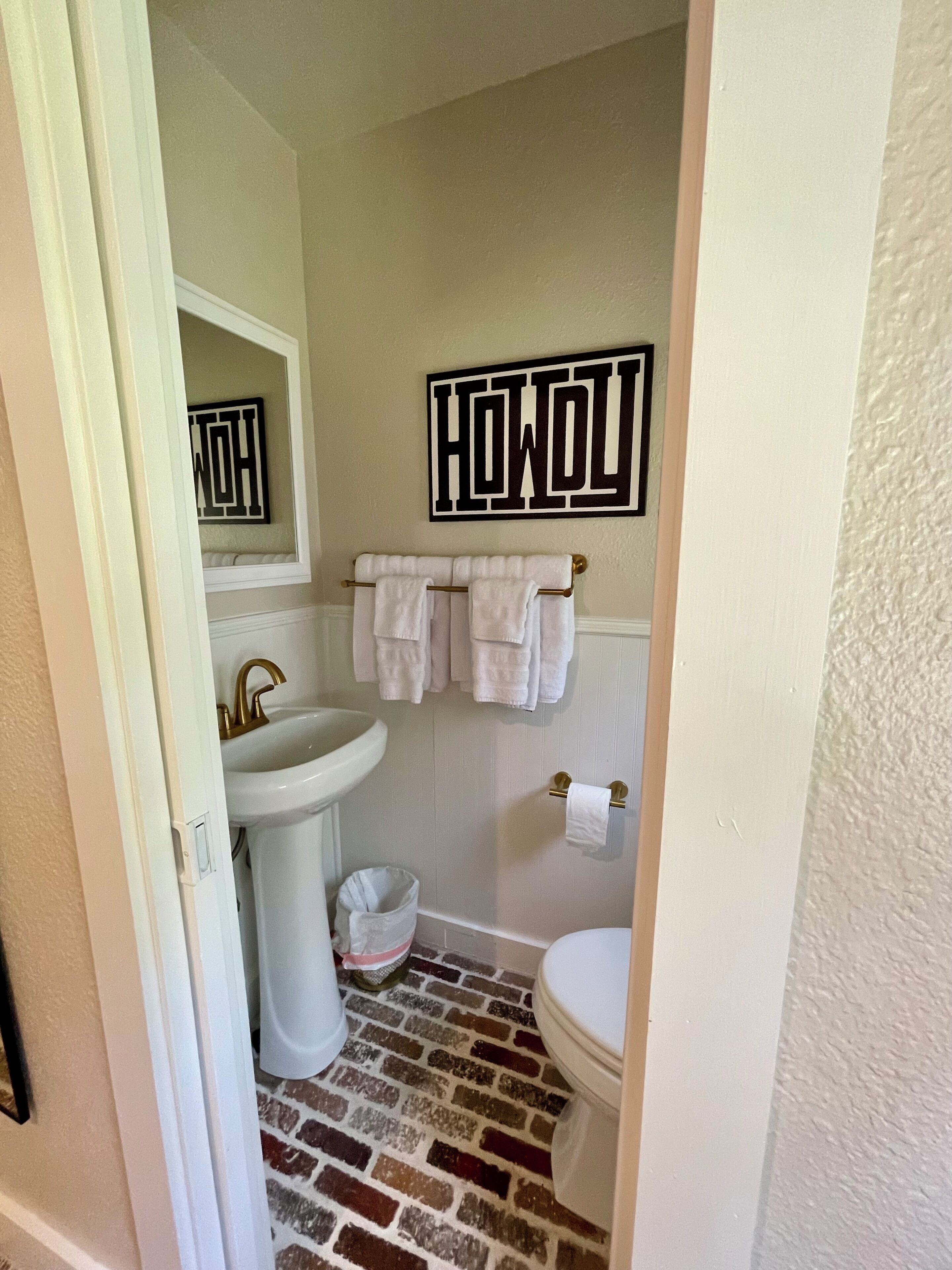 A bathroom with a white sink, a toilet, and a towel rack with multiple towels. There is a wall-mounted mirror above the sink and a decorative sign with the word 'HOWL' on the wall.
