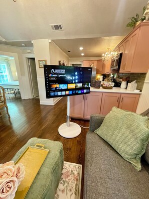 A flat-screen TV mounted on a stand in a living room with a wooden floor, a green sofa, and a floral-patterned rug. - Charming 3-bedroom cottage in Historic McKinney, three blocks from the square. (McKinney)