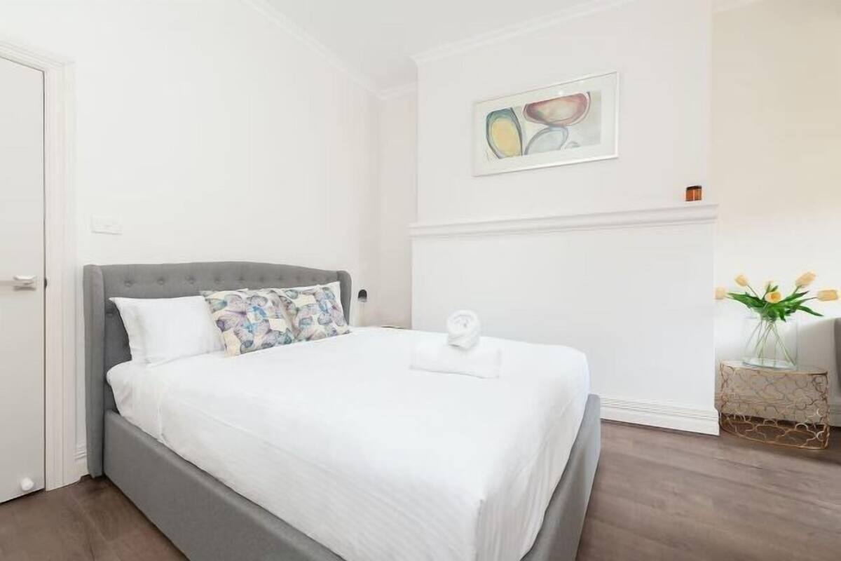 Uma cama bem arrumada com roupa de cama branca e cabeceira cinza, uma pequena mesa de cabeceira com um vaso de flores e um quadro abstrato na parede.