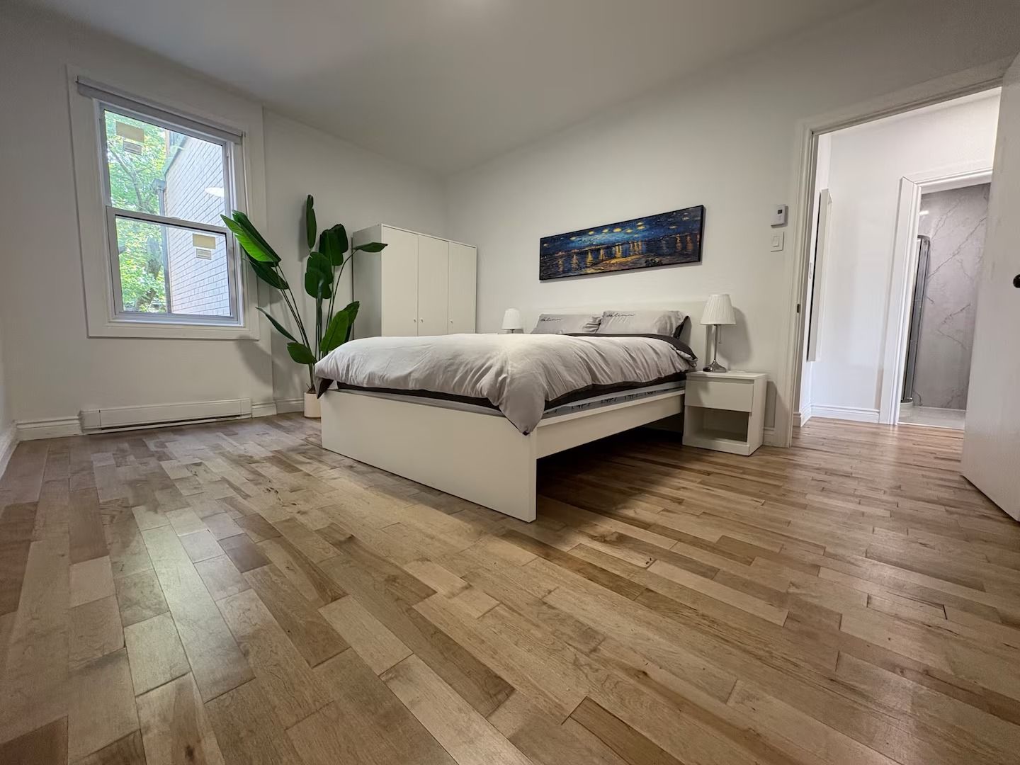 A bedroom with a bed, bedside tables, a nightstand, a potted plant, a painting, and a window.