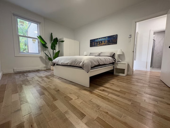 A bedroom with a bed, bedside tables, a nightstand, a potted plant, a painting, and a window.