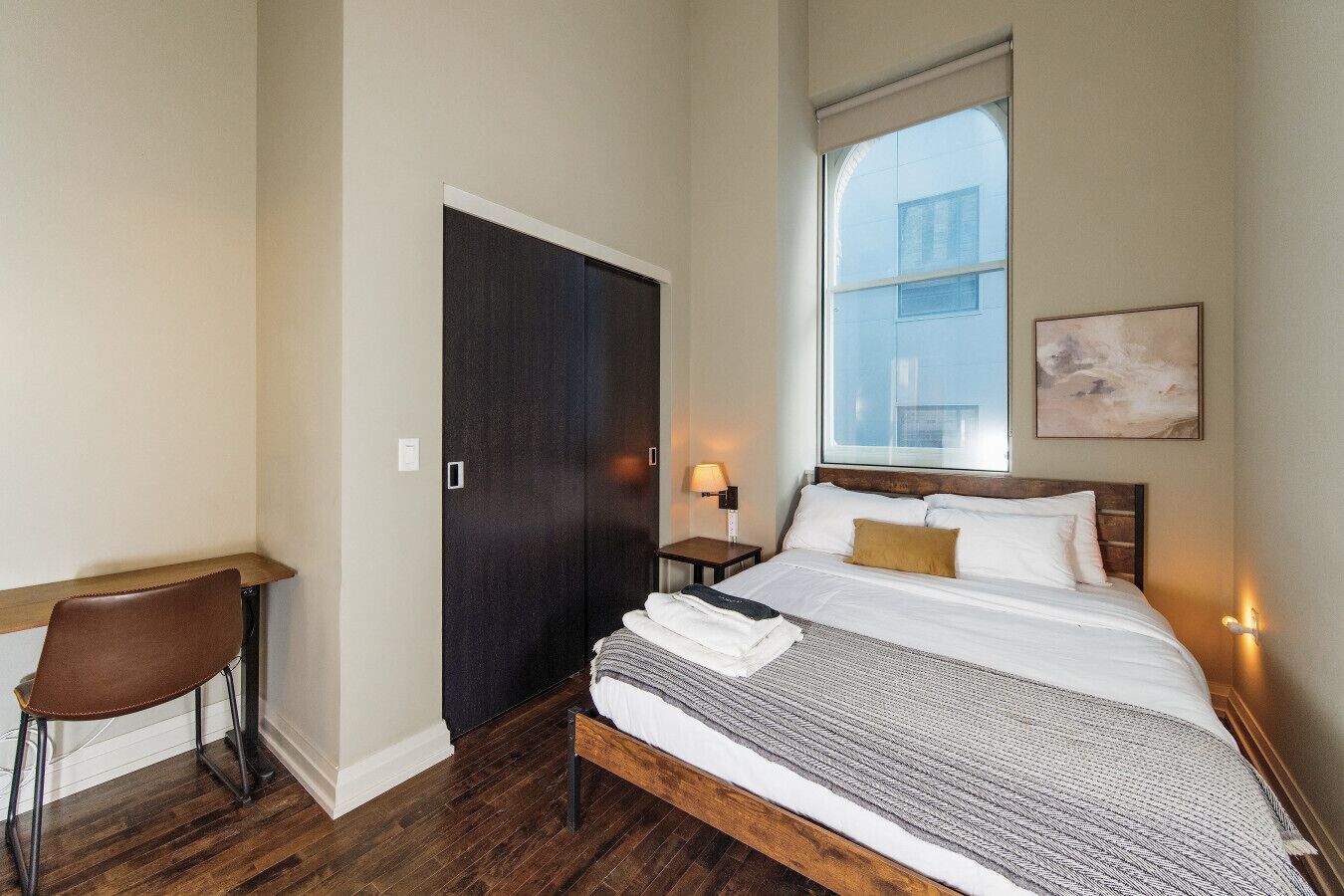 Een hotelkamer met een bed, een bureau, een stoel, een garderobe, een nachtkastje en een raam met uitzicht op een gebouw.