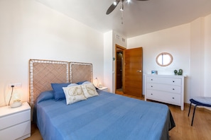 Une chambre à coucher comprenant un lit, des tables de chevet, une commode, une chaise, un ventilateur de plafond et une porte.