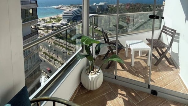 Varanda com vista para a cidade e o oceano, uma cadeira branca, uma cadeira de madeira e um vaso com planta.