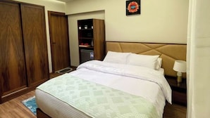 Quarto de hotel com cama, guarda-roupa de madeira, pequena escrivaninha com luminária e um quadro na parede.