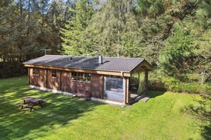 Une cabane en bois avec un toit en pente, une véranda et une table de pique-nique, située dans une zone boisée.