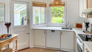 A modern kitchen with white cabinetry, a built-in dishwasher, a sink, and a stove. There are two windows with bamboo blinds, and a small table with a fruit bowl and a vase of flowers. - Awesome home in Humble with WiFi (Humble)