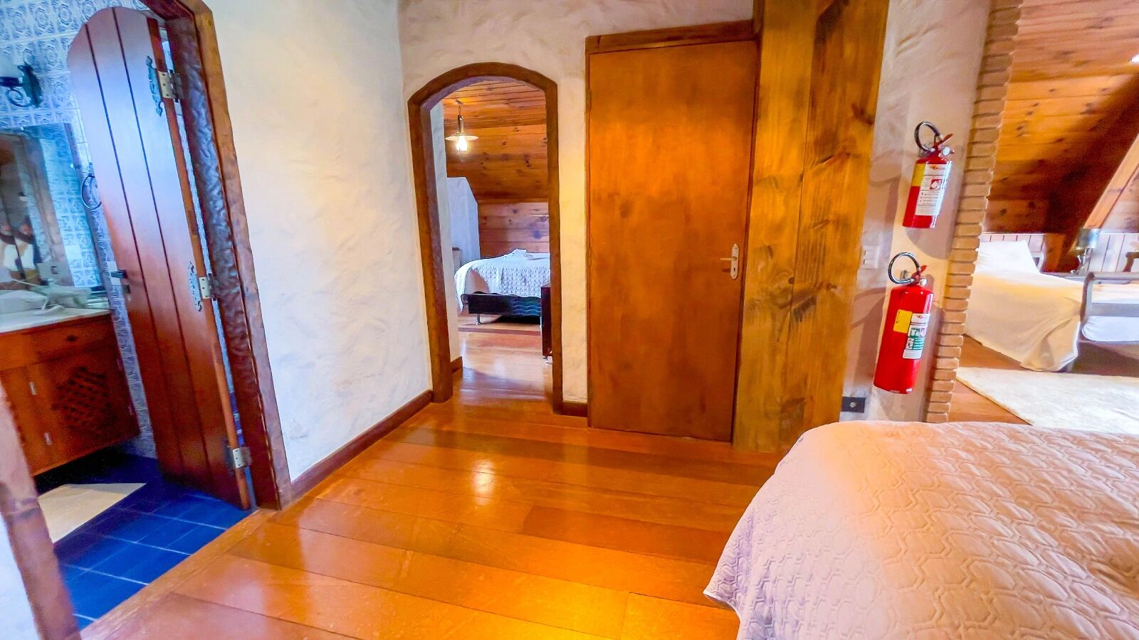 A wooden-floored hallway leading to a bedroom with a bed and a fire extinguisher mounted on the wall.