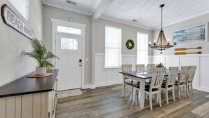 A dining area with a wooden table and chairs, a chandelier, and a wall-mounted sign reading "BEACH HOUSE". - Beachside 4BR Home • Walk to Boardwalk & Atlantic Park (Virginia Beach)