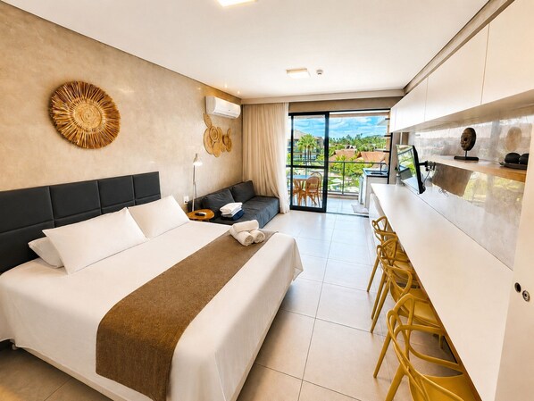 A hotel room with a bed, a sofa, a dining area with yellow chairs, a kitchen area, and a view of the outdoors.