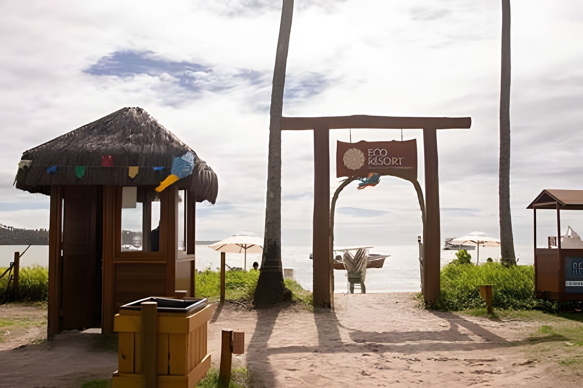 Una entrada a la playa con un arco de madera, una cabaña de techo de paja y un letrero que dice 'Eco Resort'.