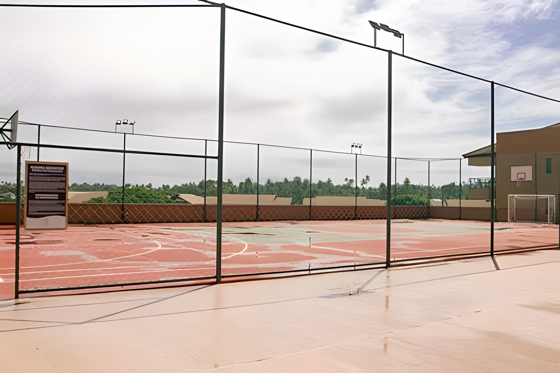 An outdoor basketball court with a metal fence, a basketball hoop, and a scoreboard.