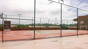 An outdoor basketball court with a metal fence, a basketball hoop, and a scoreboard.