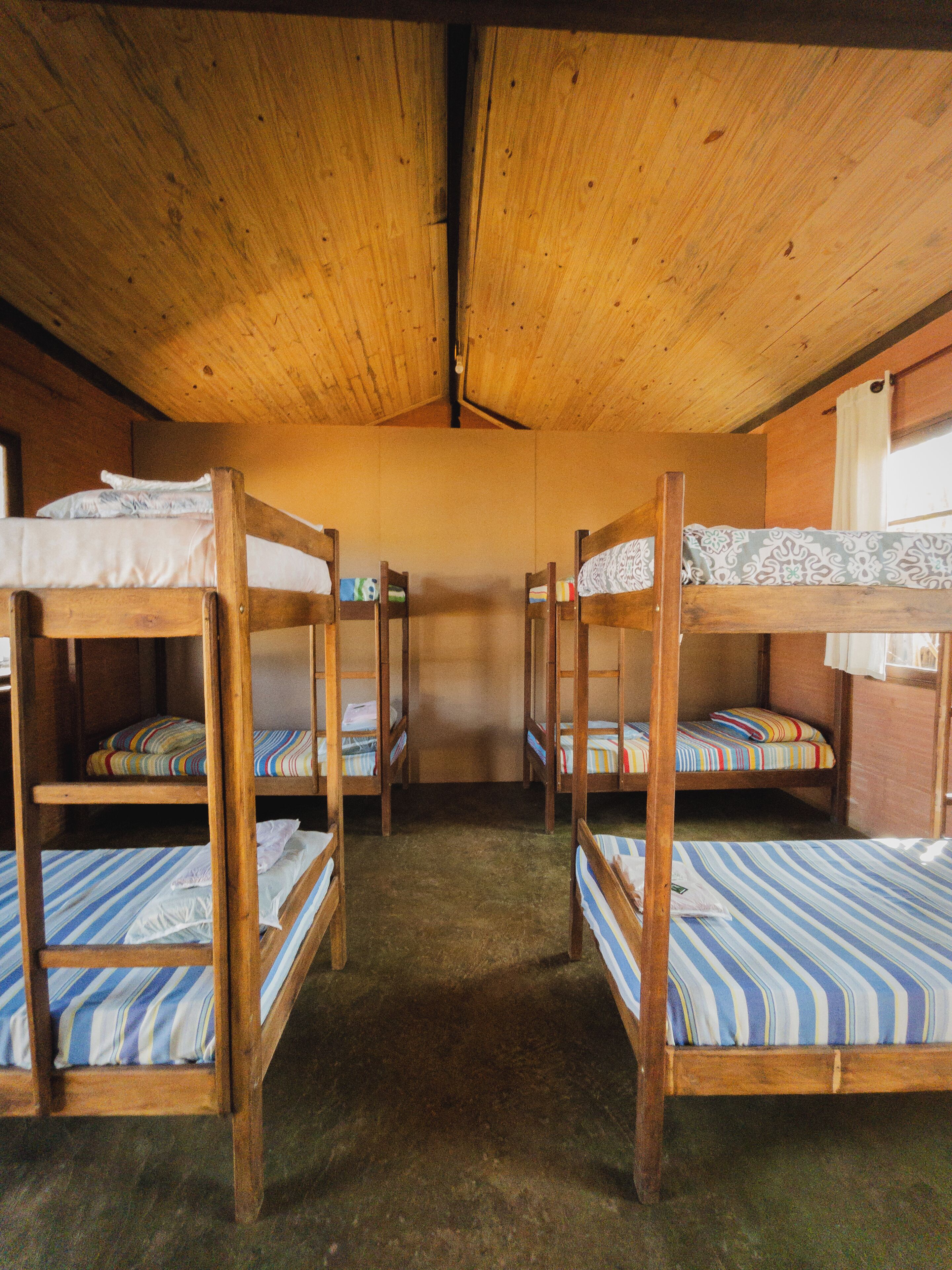 A room with four wooden bunk beds, each with a striped mattress. The room has a wooden ceiling and walls, and a carpeted floor.
