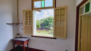 A room with a window featuring wooden shutters, a small table with a vase, and a view of greenery outside. - Camping Tiradentes (Tiradentes)