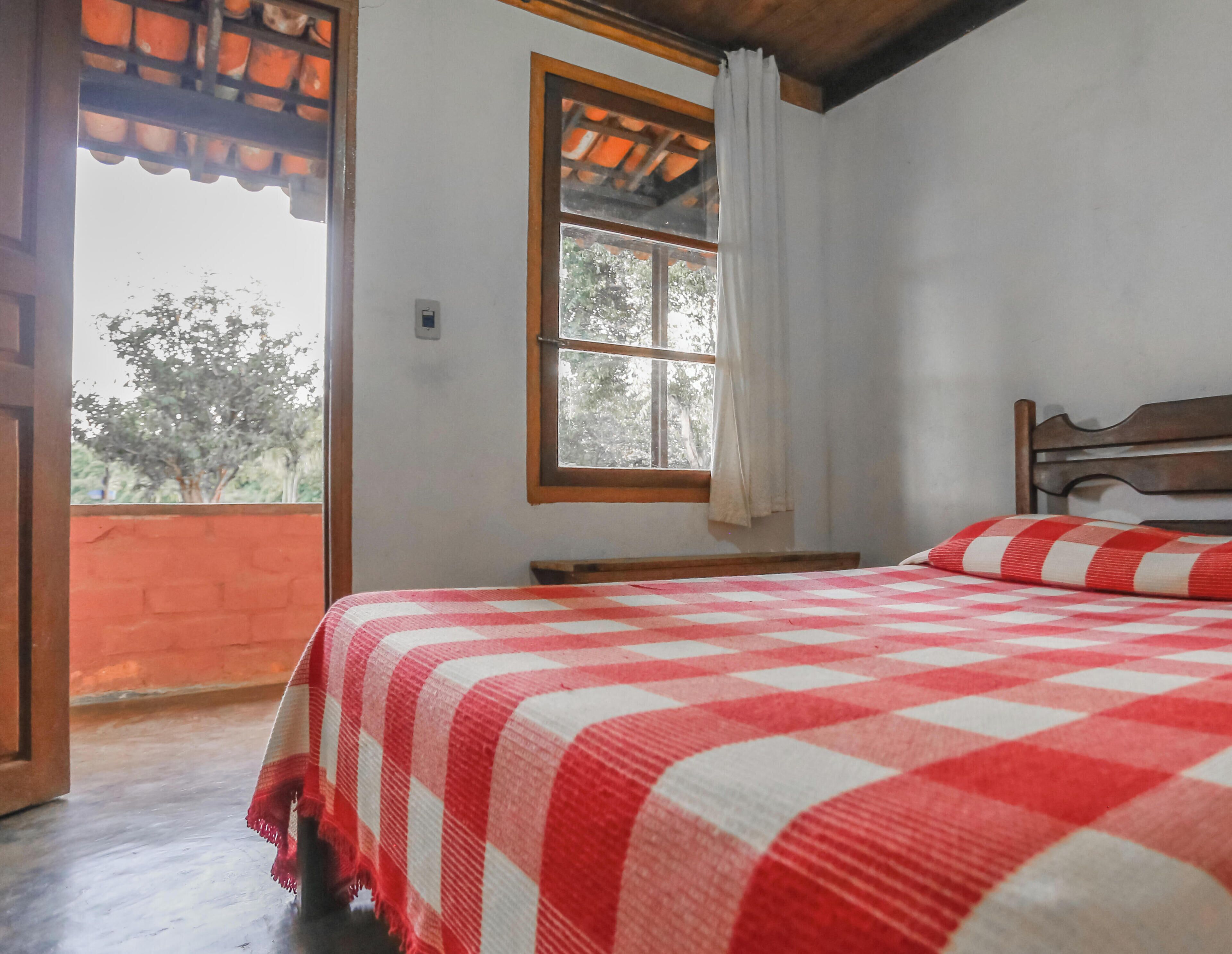 A bedroom with a checkered bedspread, a wooden headboard, a window with a curtain, and a view of trees outside.