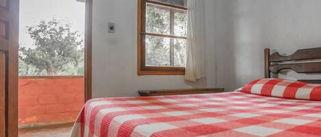 A bedroom with a checkered bedspread, a wooden headboard, a window with a curtain, and a view of trees outside.